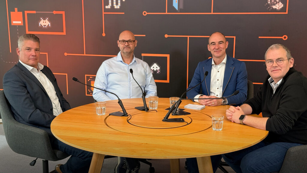 Four guests at a round wooden table with table microphones in front of an orange wall